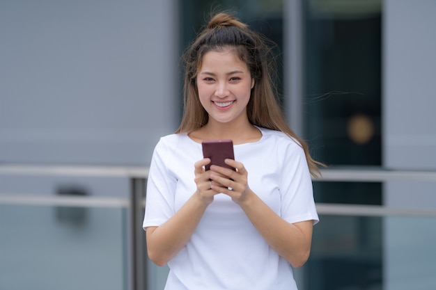 Mujer en camiseta blanca y jeans usando teléfono de pie en un piso afuera en el fondo de la calle de la ciudad, día de verano