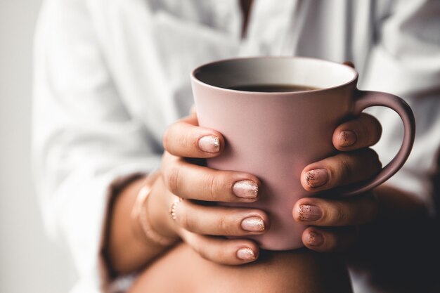 Mujer con una camiseta blanca sostiene el café de la mañana en una taza de cerámica rosa. Manicura. Vista frontal