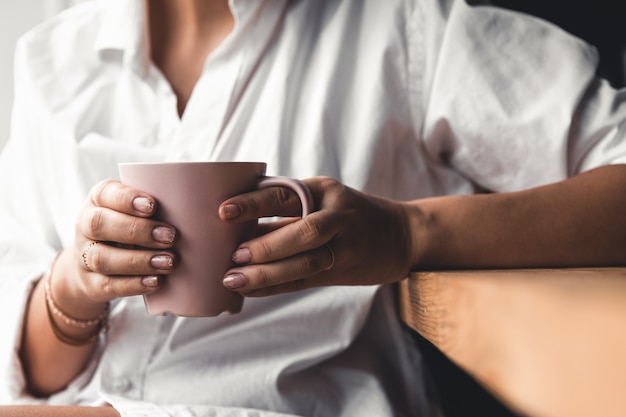 Mujer con una camiseta blanca sostiene el café de la mañana en una taza de cerámica rosa. Manicura. Vista frontal