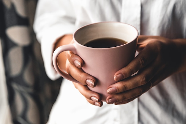 Mujer con una camiseta blanca sostiene el café de la mañana en una taza de cerámica rosa. Manicura. Vista frontal