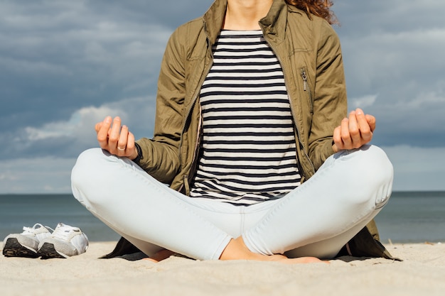 La mujer con camiseta a rayas y el abrigo verde está sentada en postura de meditación en la playa