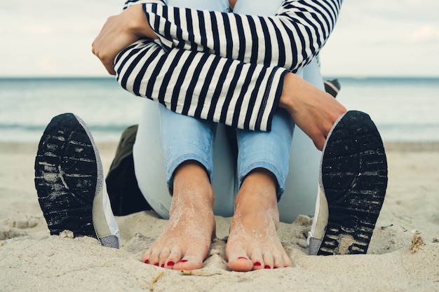 Mujer en camiseta a rayas y pantalones vaqueros se sienta descalza en la playa al lado de los zapatos