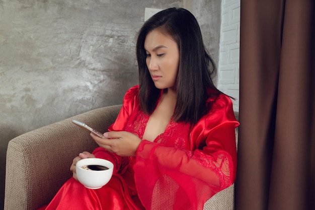 Mujer en camisón rojo sosteniendo una taza de café y mirando un teléfono inteligente