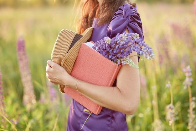 Mujer en el campo de puesta de sol de verano con ramo de flores silvestres sombrero de altramuces y cuaderno rosa Cercanía al concepto de autodescubrimiento de la naturaleza Concepto de relajación y bienestar Concepto de buen humor de día soleado