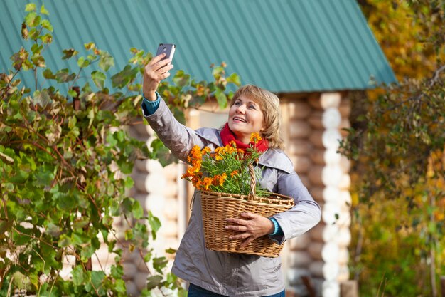 Una mujer con una canasta está en el jardín. Una mujer de mediana edad en jeans y una chaqueta sostiene flores contra los árboles en otoño. Ella esta sosteniendo el telefono