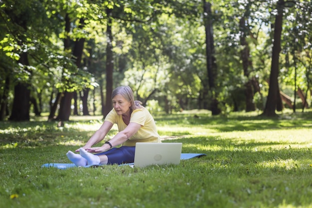 Mujer canosa activa en el parque haciendo ejercicio sentada en la alfombra y el césped usando una computadora portátil para clases en línea en el parque