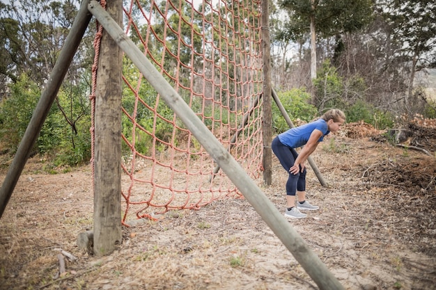 Mujer cansada agacharse con las manos en las rodillas durante la carrera de obstáculos en el campo de entrenamiento