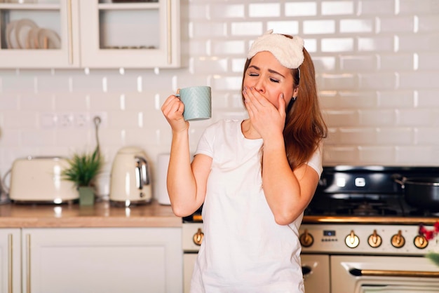 Mujer cansada y dulce sosteniendo una taza de café en la cocina de casa, bostezos. Chica, la mujer no durmió lo suficiente