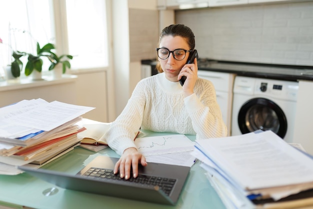 Una mujer cansada con gafas hace una pausa de su trabajo remoto hablando por teléfono en medio de una mesa