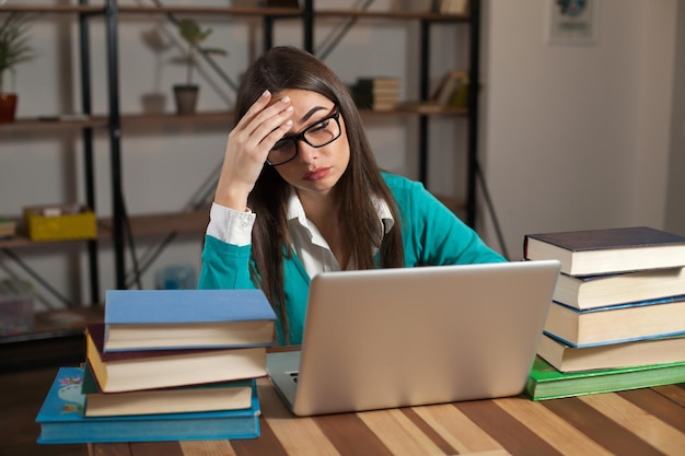 Una mujer cansada con gafas con muchos libros y una computadora portátil está sentada en la mesa