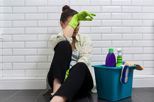 Mujer cansada haciendo limpieza de la casa en el baño. Mujer en guantes con detergentes, cubo sentado en el suelo. Fatiga, estrés, limpieza, hogar