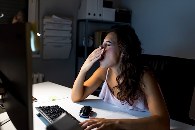 Mujer cansada con papeles y laptop bostezando en la oficina de noche