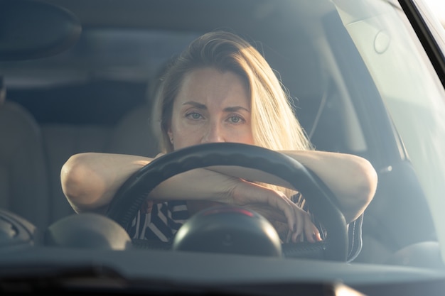 Mujer cansada pensativa conduciendo un coche mirando a través del parabrisas a la cámara pensando en algo