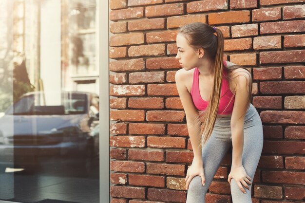 Mujer cansada recostada sobre la pared de ladrillo, relajándose del entrenamiento de jog, espacio de copia