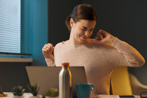 Foto mujer cansada sentada en el escritorio y trabajando con su computadora portátil está estirando los brazos y la espalda