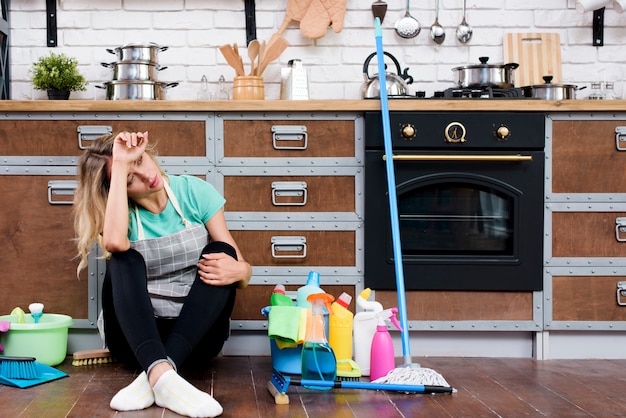 Mujer cansada sentada en el piso de la cocina con productos y equipos de limpieza