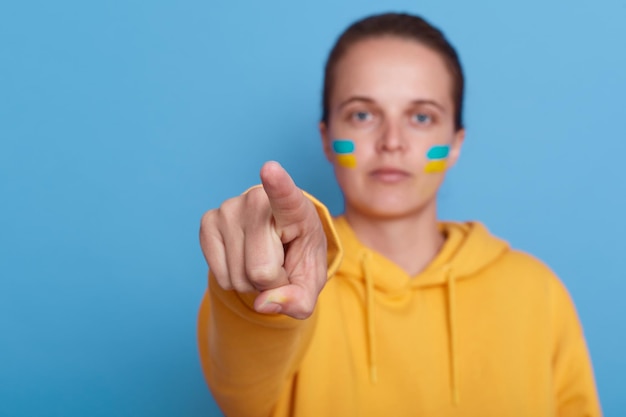 Mujer con capucha amarilla y con la bandera de Ucrania en la mejilla aislada sobre fondo azul mujer señalando con el dedo a la cámara Dejar de luchar en Ucrania Rusia detener la guerra
