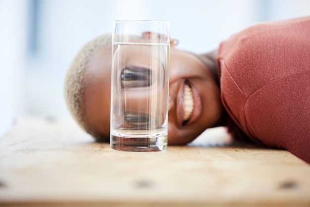 Mujer cara graciosa y vaso de agua con grandes ojos riendo y broma cómica en la mesa en el apartamento Chica africana loca joven estudiante y comedia para meme con sonrisa feliz en casa y jugando en el escritorio