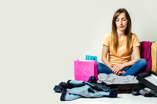 Mujer con una cara triste sentada en el suelo rodeada de diferentes conjuntos y bolsas de compras.