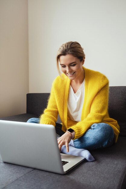 Mujer con cárdigan amarillo sentada en el sofá y usando una computadora portátil