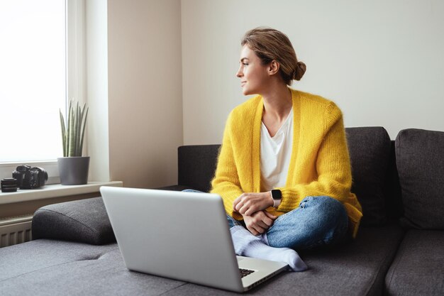 Mujer con cárdigan amarillo sentada en el sofá y usando una computadora portátil