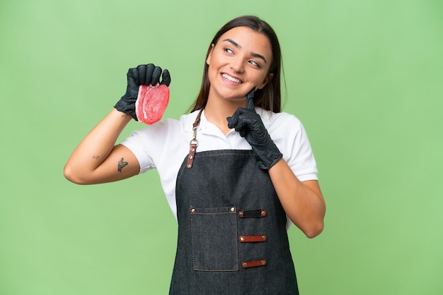 Mujer carnicera con un delantal y sirviendo carne fresca cortada aislada en un fondo de croma verde pensando en una idea mientras mira hacia arriba