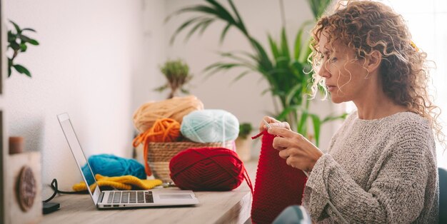 Mujer en casa en actividades de ocio aprendiendo de clases en línea para tejer trabajo Mujeres usando laptop para su hobby o trabajo alternativo Señora de mediana edad tejiendo con lana roja Concepto de usar web