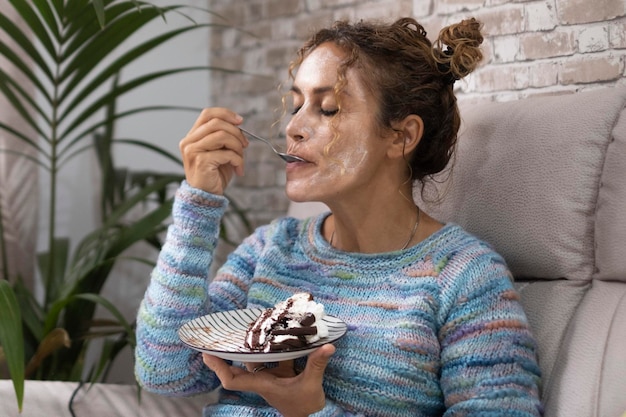 Una mujer en casa disfrutando de comer un sabroso pastel con crema y chocolate con expresión de placer en la cara Gente femenina relajándose en casa Concepto de comida y felicidad Joven adulta en el sofá