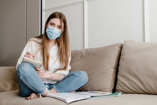 Mujer en casa en el sofá con máscara estudiando con libros y cuaderno