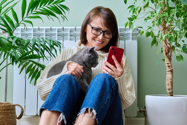 Mujer en casa usando un teléfono inteligente calentándose con un gato cerca del radiador de calefacción