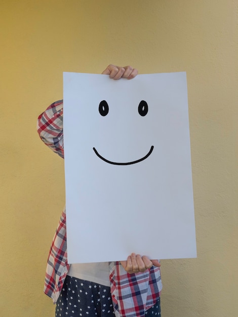 Mujer casual escondiendo su rostro detrás de una valla publicitaria en blanco con una cara sonriente Copia espacio