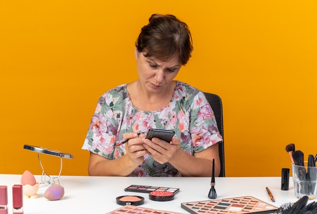 Mujer caucásica adulta despistada sentada a la mesa con herramientas de maquillaje sosteniendo pincel de maquillaje y mirando el teléfono aislado en la pared naranja con espacio de copia