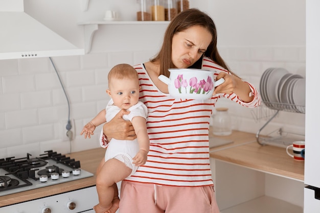 Mujer caucásica adulta joven parada en la mesa en la cocina y oliendo comida en una olla mientras habla por teléfono móvil y sostiene a su hija pequeña en las manos