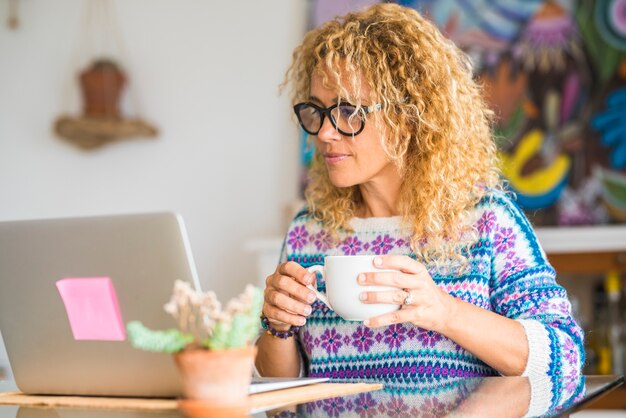 Mujer caucásica adulta mirar computadora portátil en casa bebiendo con una taza - gente bonita rubia usa tecnología para el trabajo o el ocio - joven conectada con gafas negras