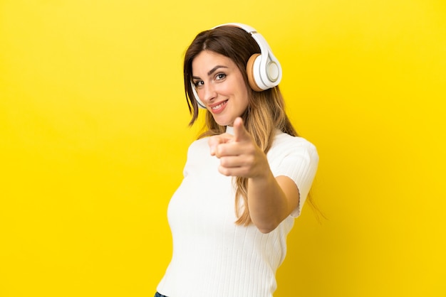 Mujer caucásica aislada sobre fondo amarillo escuchando música y apuntando hacia el frente