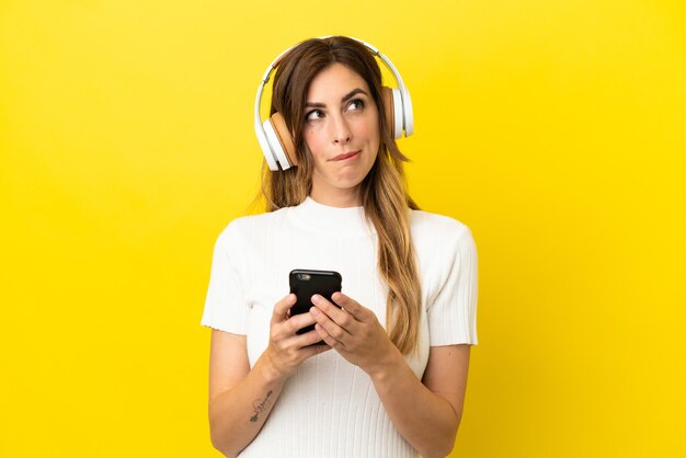 Mujer caucásica aislada sobre fondo amarillo escuchando música con un móvil y pensando