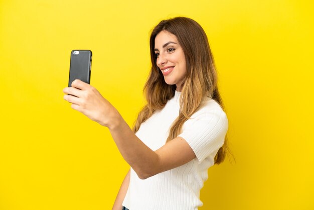 Mujer caucásica aislada sobre fondo amarillo haciendo un selfie con teléfono móvil