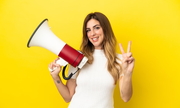 Mujer caucásica aislada sobre fondo amarillo sosteniendo un megáfono y sonriendo y mostrando el signo de la victoria