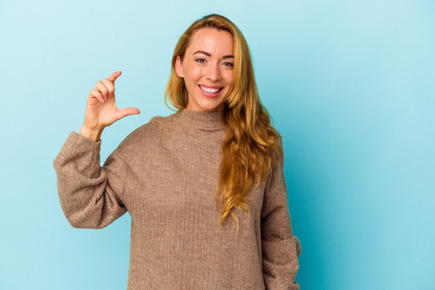 Mujer caucásica aislada sobre fondo azul sosteniendo algo poco con los dedos índices, sonriendo y confiado.