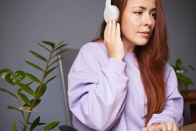 Mujer caucásica alegre escuchando música o podcast y sosteniendo un teléfono inteligente Muchas plantas verdes alrededor Mujer pelirroja con suéter violeta