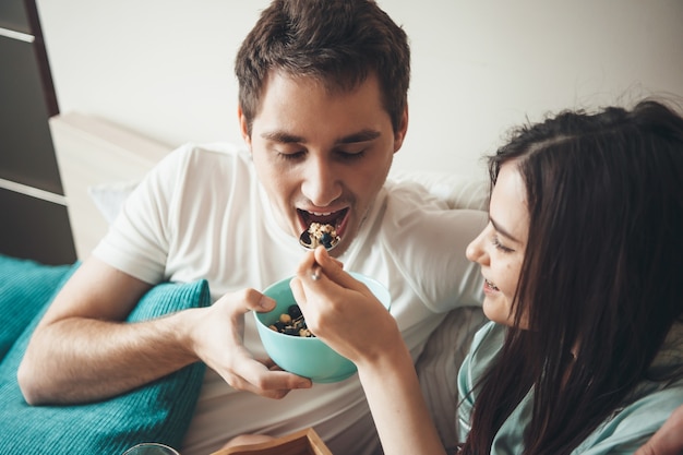 Mujer caucásica alimentando a su amante con cereales en la cama está sonriendo mientras lo mira