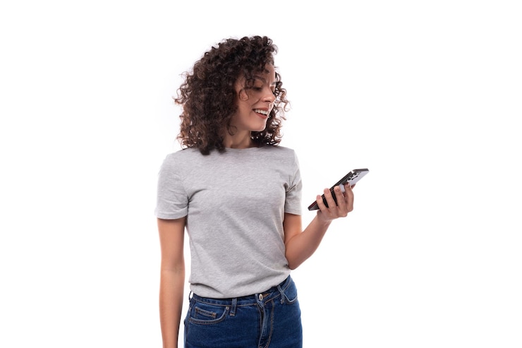 Foto mujer caucásica de un año de edad agradable y delgada con cabello rizado en una camiseta gris sostiene un teléfono inteligente
