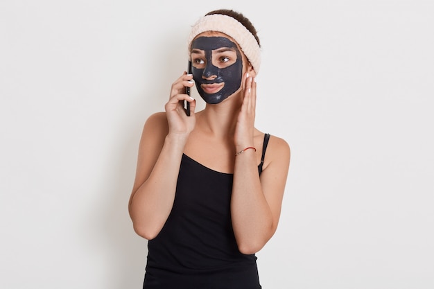 Mujer caucásica con banda para el cabello en la cabeza y mascarilla negra hablando con su amiga usando un teléfono móvil aislado sobre la pared blanca, mirando a un lado.
