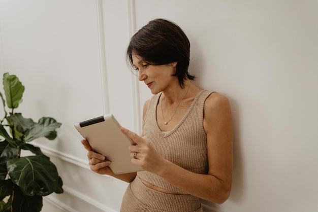 Mujer caucásica bastante adulta mira cuidadosamente la pantalla de la tableta en sus manos mientras está de pie contra la pared blanca Concepto de compras en línea