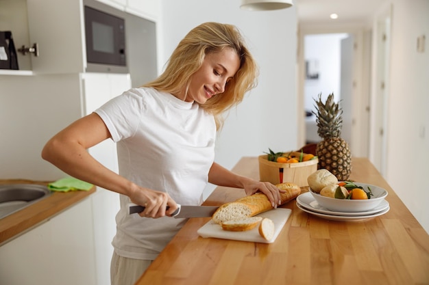 Mujer caucásica bastante joven cortando diapositivas de baguette en la cocina por la mañana