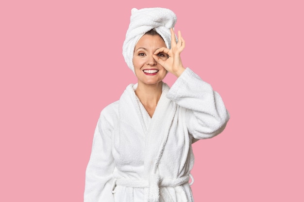 Mujer caucásica en bata de baño y toalla emocionada manteniendo el gesto ok en el ojo