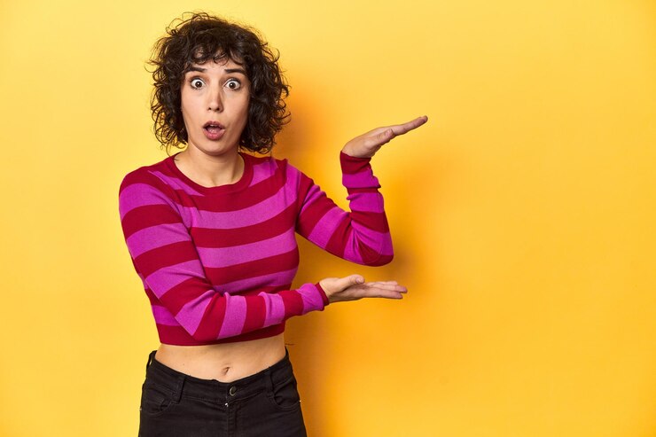 Foto mujer caucásica de cabello rizado con camiseta a rayas rosas conmocionada y asombrada sosteniendo un espacio de copia entre las manos