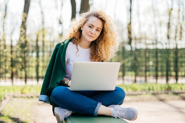 Mujer caucásica con cabello rizado claro y ojos azules con ropa cómoda sentada con las piernas cruzadas al aire libre sosteniendo una laptop de rodillas enviando mensajes con su novio mirando directamente a la cámara