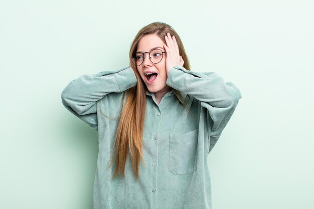 Mujer caucásica de cabello rojo que se siente feliz emocionada y sorprendida mirando hacia un lado con ambas manos en la cara