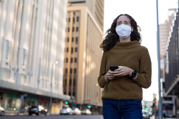 Foto mujer caucásica en las calles de la ciudad durante el día, usando una máscara facial contra el coronavirus covid19 de pie y usando su teléfono inteligente